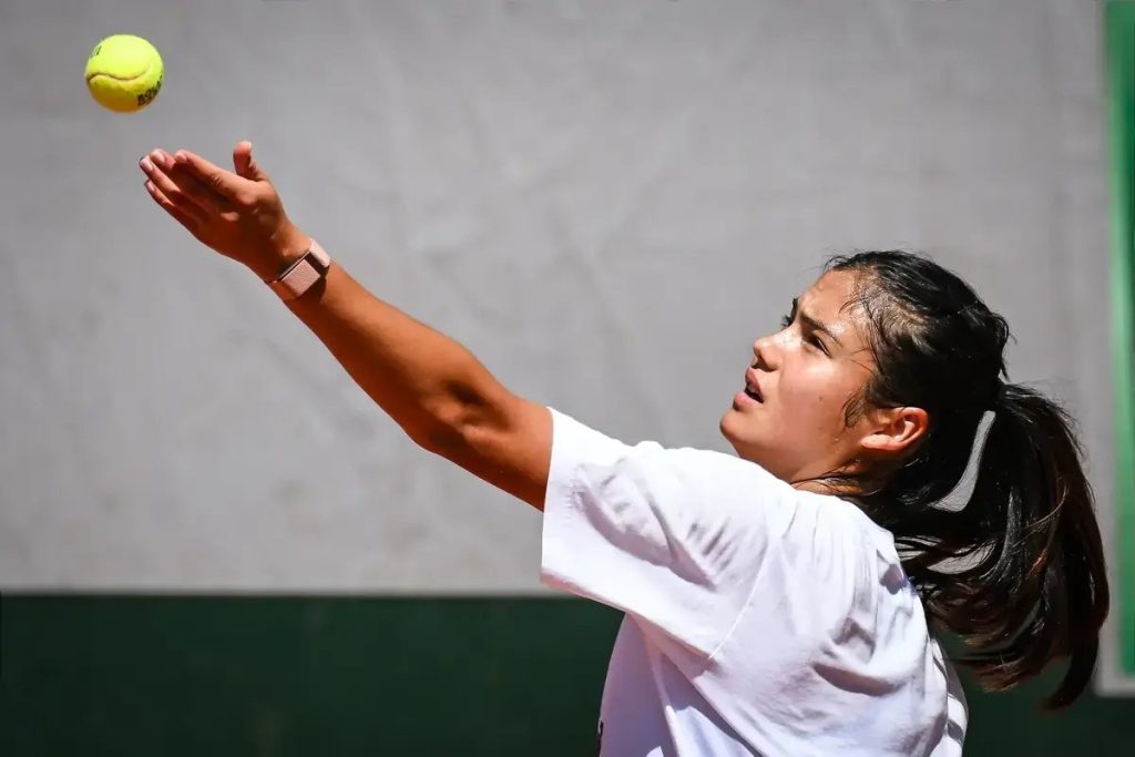 A tennis player in a white shirt tossing a yellow ball overhead to serve on a clay court, with a green backdrop