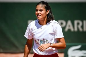 A tennis player in a white graphic t-shirt and red skirt on a green court at Roland-Garros, with Lacoste and PARIS signage behind her.