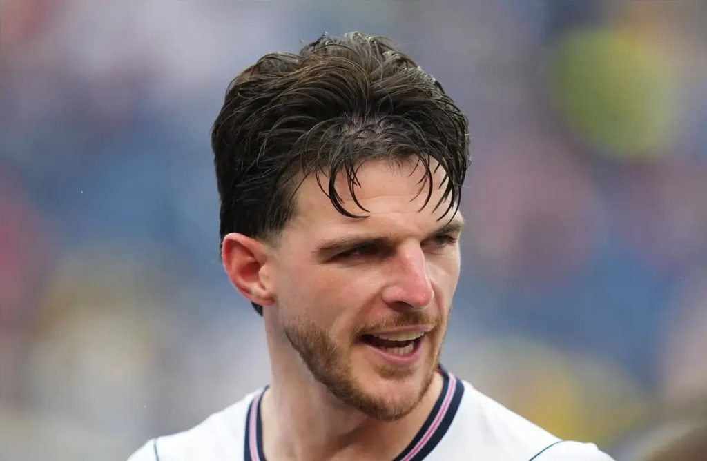A footballer with swept-back dark hair and stubble, wearing a white kit with navy and pink trim, speaking on a stadium pitch.