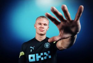 A footballer in a black Manchester City training kit with the number 9 and OKX sponsor, holding up a hand toward the camera against a blue backdrop