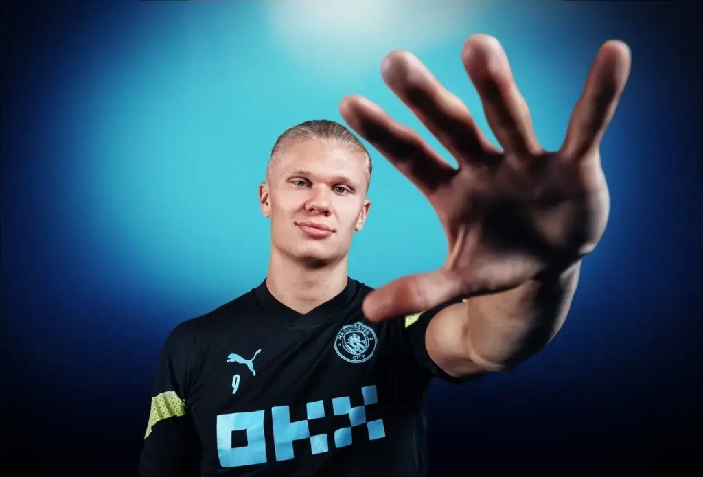 A footballer in a black Manchester City training kit with the number 9 and OKX sponsor, holding up a hand toward the camera against a blue backdrop
