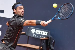 Tennis player in black shirt and blue headband hitting a backhand with a blue Babolat racket, emu sign behind him at a clay court event