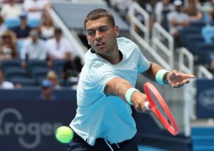 Tennis player in a light blue shirt and green wristbands hitting a forehand with a red racket at a hard-court event, spectators behind him.