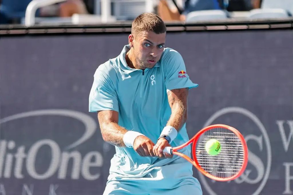 Tennis player in a light blue On shirt with Red Bull logo hits a backhand return with a red racket at a Cincinnati event court.