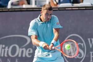 Tennis player in a light blue On shirt with Red Bull logo hits a backhand return with a red racket at a Cincinnati event court.