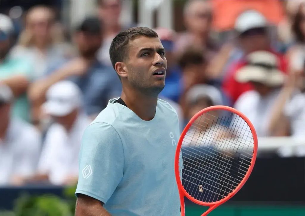 Tennis player in a light blue shirt holding a red racquet, looking up during a match with spectators blurred behind him