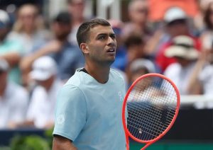 Tennis player in a light blue shirt holding a red racquet, looking up during a match with spectators blurred behind him