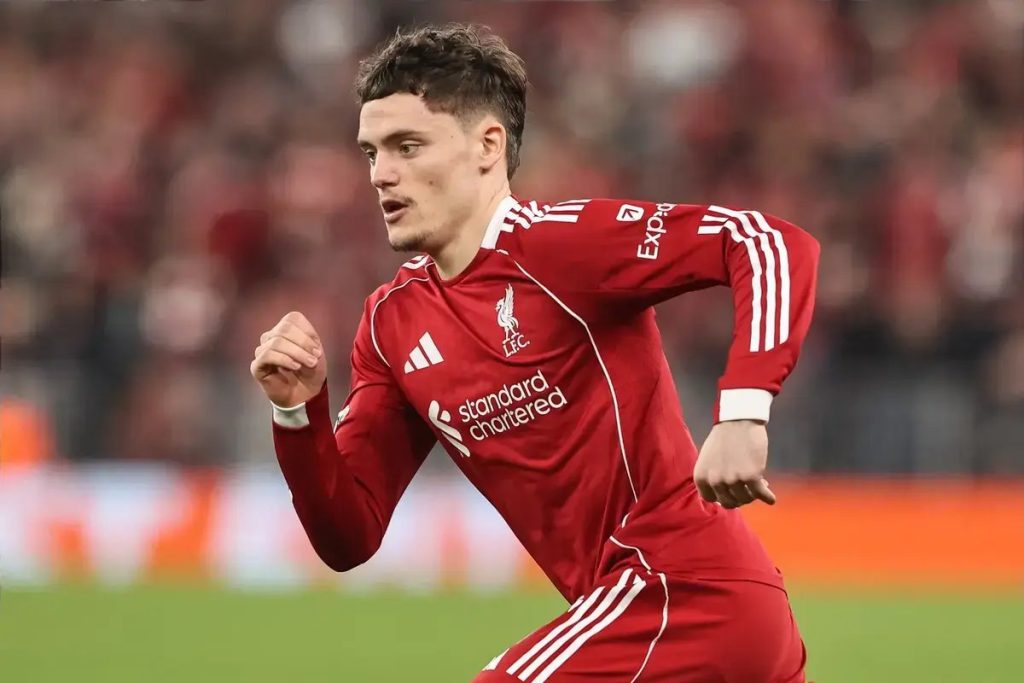 Young male footballer in a red Liverpool FC kit with Standard Chartered sponsor, running during a match in a blurred stadium setting