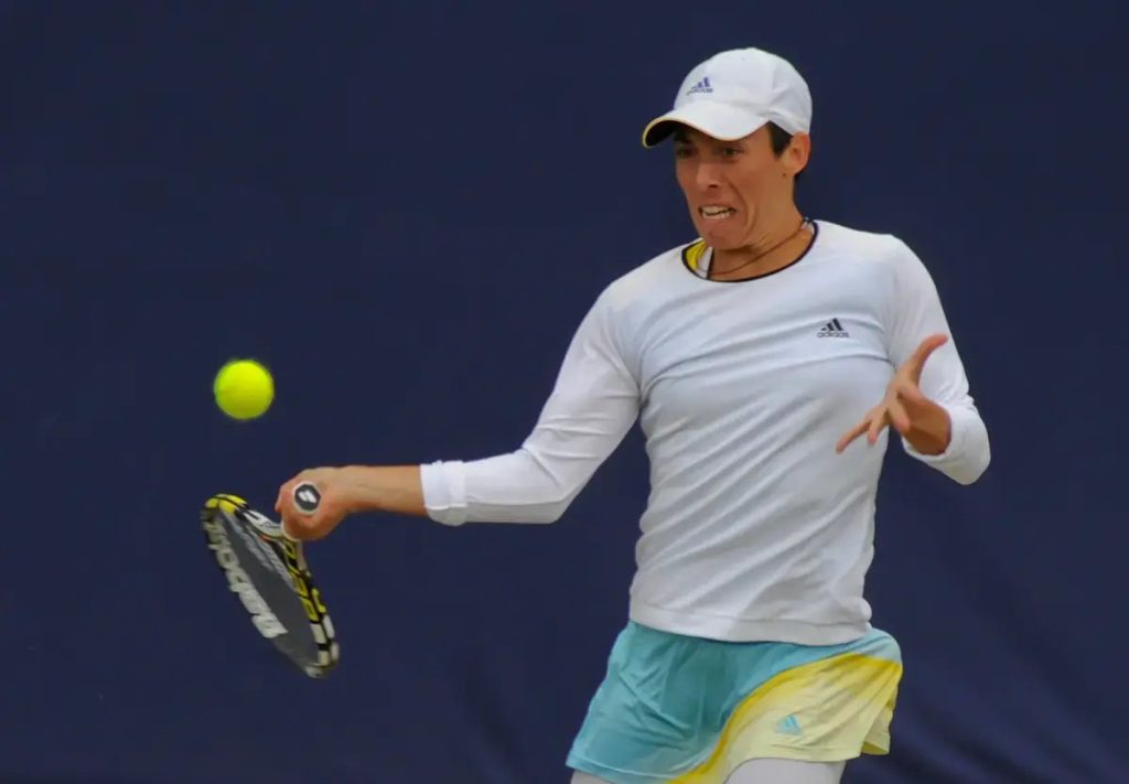 A tennis player in a white Adidas cap and long-sleeve top swings a forehand on a blue court, ball in view.