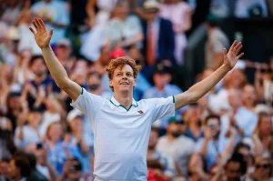 A tennis player with curly auburn hair in a white polo, arms raised, celebrating before a cheering crowd at a grass-court tournament.