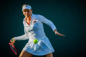 A tennis player in a white visor and white ellesse dress hits a forehand with a red racket, a yellow ball in motion, on a dark court