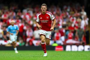 Arsenal player in red and white home kit, number 14, running on the pitch during a match with a blurred crowd behind him.