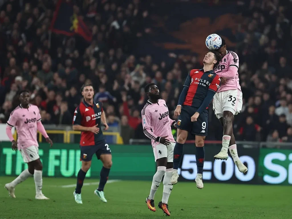 Jogadores do Genoa de azul e vermelho e do Juventus de rosa disputam uma bola alta, o número 9 do Genoa e o 32 do Juventus, num estádio cheio.