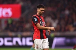 Jogador de futebol com camisola vermelha e preta do AC Milan, calção branco e número 9, em campo durante um jogo, de cabeça baixa.