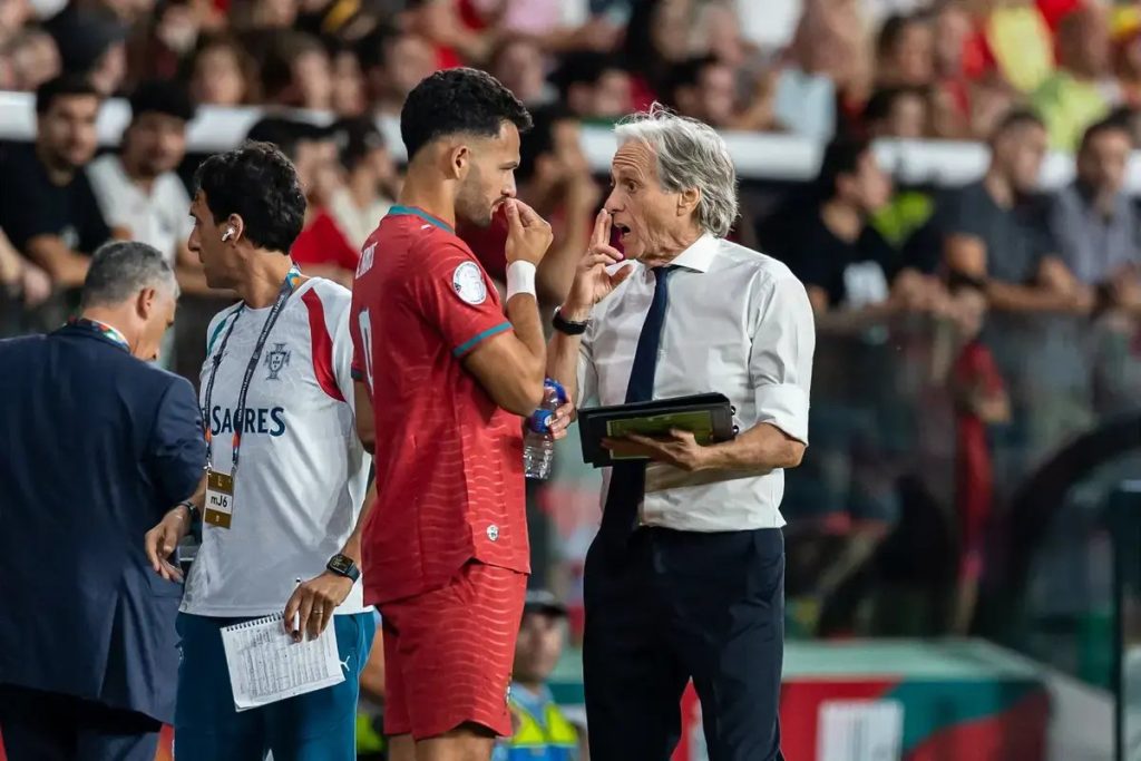 Portugal player in red kit touching his face beside a grey-haired coach in a white shirt and dark tie during a match.