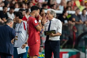 Portugal player in red kit touching his face beside a grey-haired coach in a white shirt and dark tie during a match.