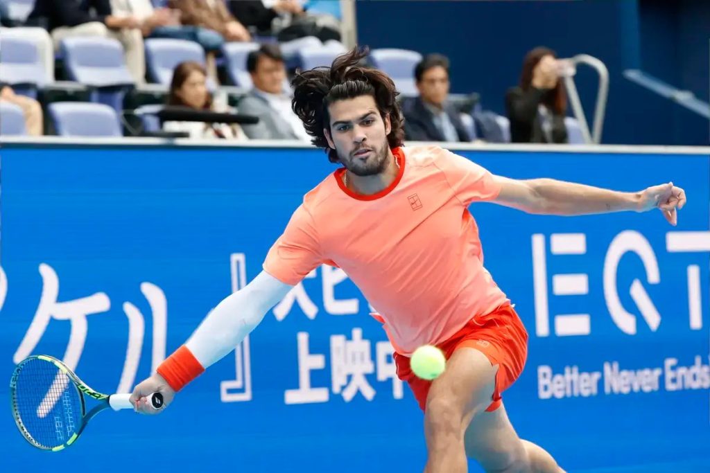 A tennis player in an orange shirt and red shorts reaches low for a forehand return during a match, with a blue "EQT" branded court wall behind him.
