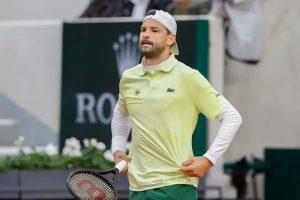 Tennis player in a light green Lacoste polo and white cap holding a Wilson racket on a clay court, with a Rolex sign behind him.