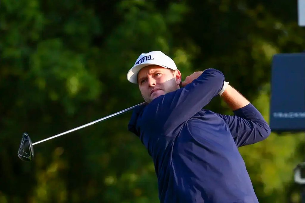 Golfer in navy long-sleeve shirt and white cap swings a driver on a tree-lined course, following through on his tee shot