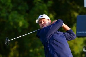 Golfer in navy long-sleeve shirt and white cap swings a driver on a tree-lined course, following through on his tee shot