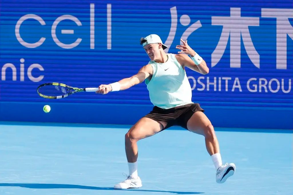 A tennis player in a mint sleeveless top and dark shorts hits a forehand on a blue court, with a Cellular-branded backdrop behind him.