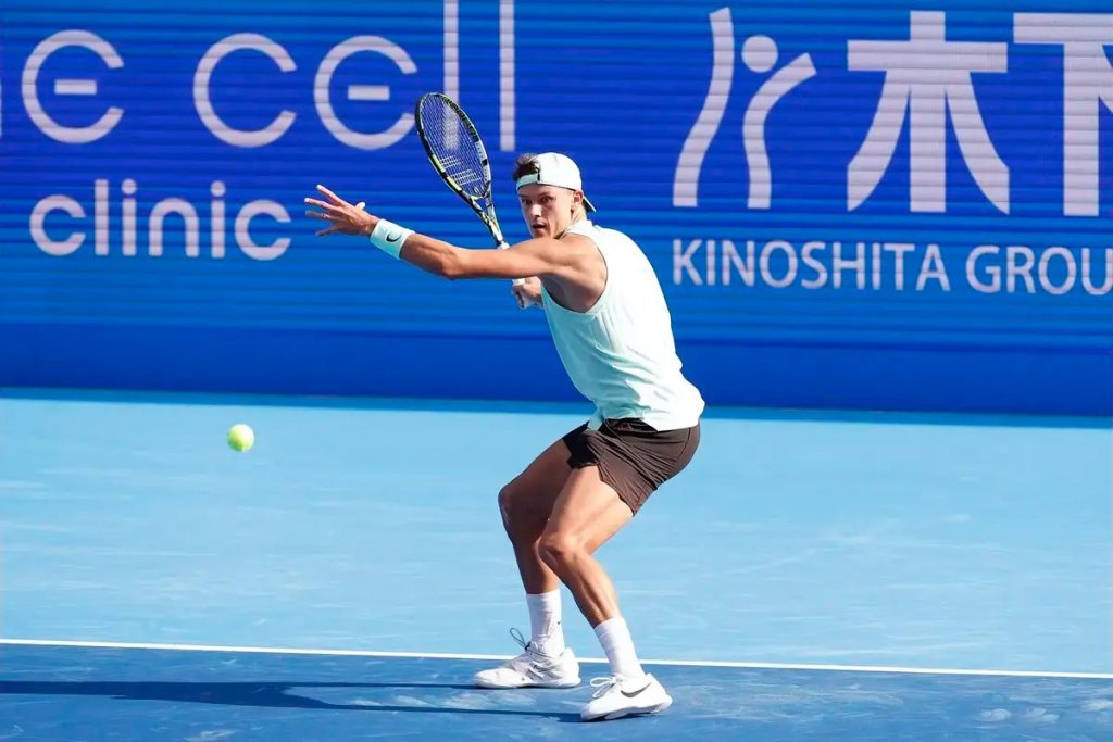 Tennis player in a white cap and sleeveless mint top hits a forehand on a blue court, sponsor boards behind him.