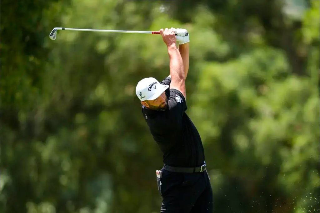 Golfer in black shirt and white Callaway cap follows through a swing on a sunny course, with blurred green trees behind him.
