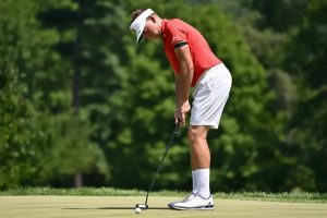 A golfer in a red polo, white visor and shorts lines up a putt on a green, with trees behind him