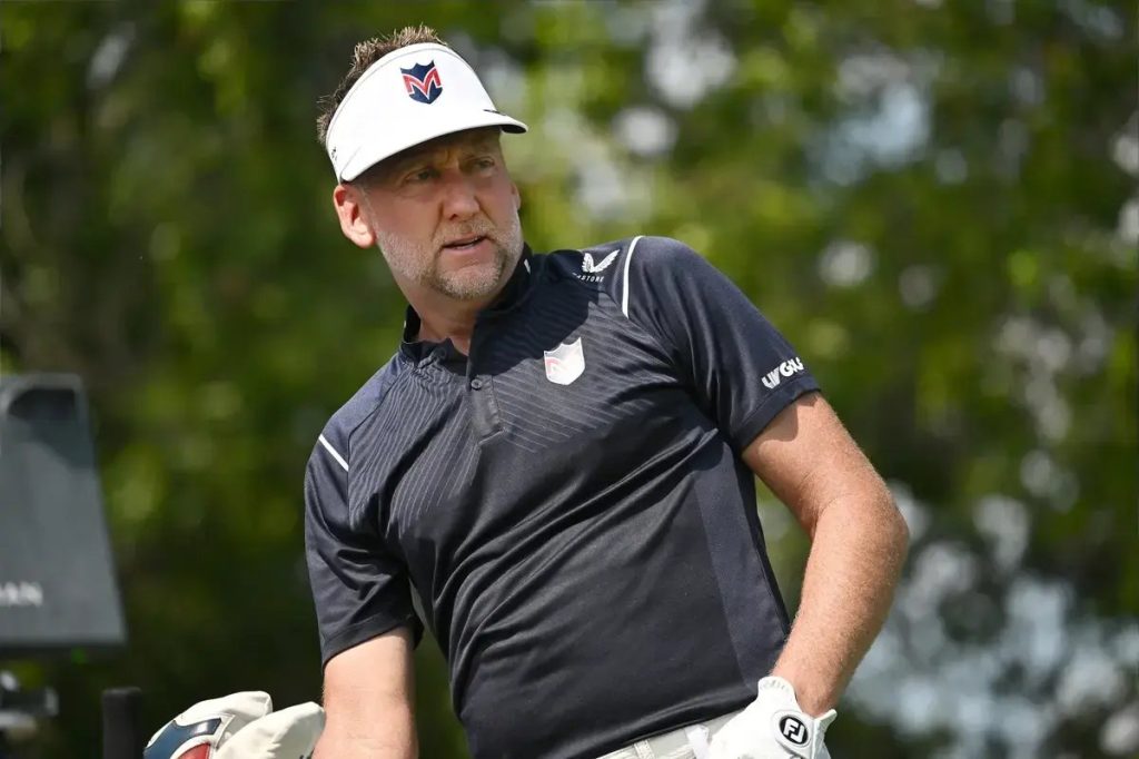 Golfer in navy polo and white visor with gloves, looking to the side on a golf course with green trees behind him
