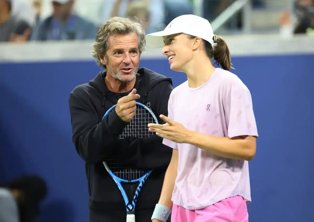 Tennis player in a white cap and pink On shirt laughing beside a grey-haired man in a black jacket holding a blue racket, on a blue court.