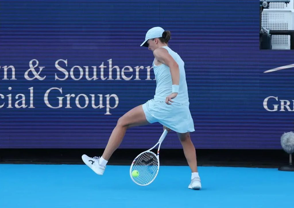 Tennis player in light blue dress and cap lifts her leg mid-swing on a blue court, racket low, ball on the ground, Southern Financial Group sign behind.