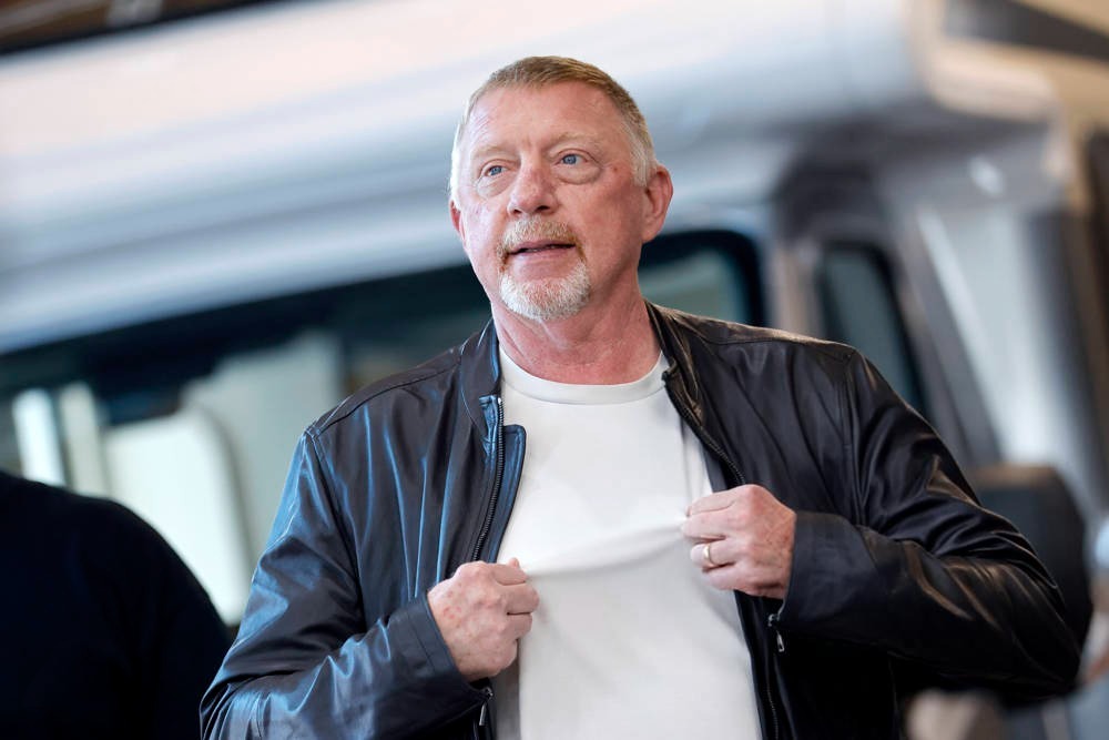 Man in dark leather jacket over white t-shirt, pulling his shirt collar at an outdoor event, with a blurred vehicle behind him.