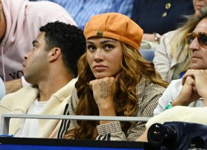 A woman in an orange cap and plaid jacket watches a match intently, chin resting on her fist, beside a man in a cream jacket at the US Open.