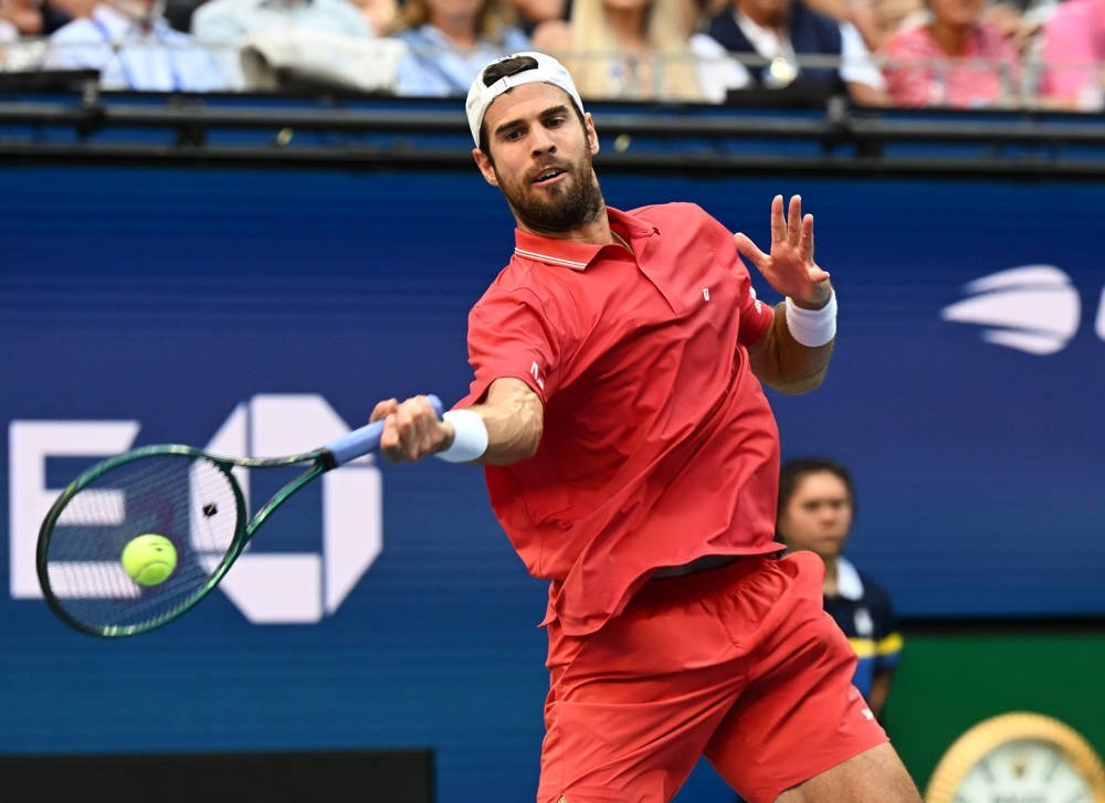 Tennis player in red shirt and white cap hitting a forehand return on a blue court, with a yellow ball in motion.