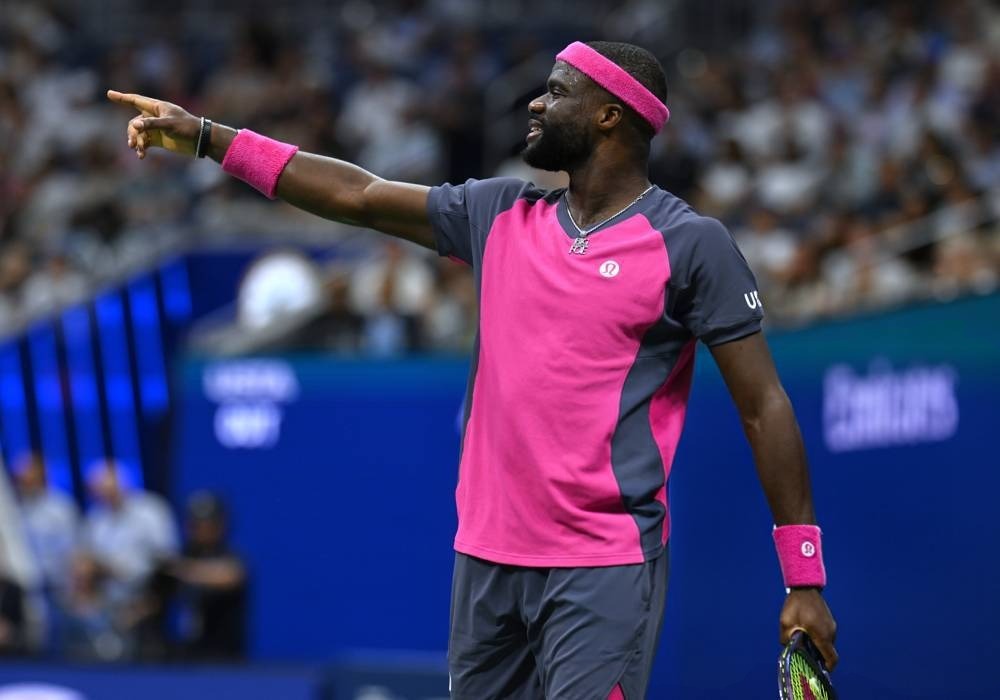 Tennis player in pink and grey kit and pink wristband points while speaking, on court at a packed stadium during the US Open