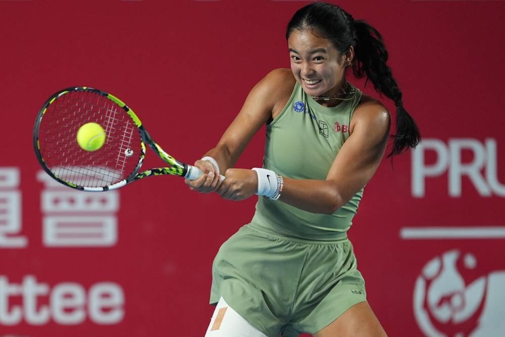 Tennis player in a sage green sleeveless outfit hitting a two-handed backhand on a red court at a tournament; Alexa Eala is the article's subject.