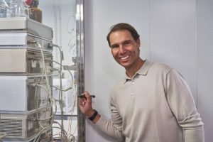 A smiling man in a beige sweater signs a glass panel beside stacked laboratory equipment with tubing, in a clean lab setting.