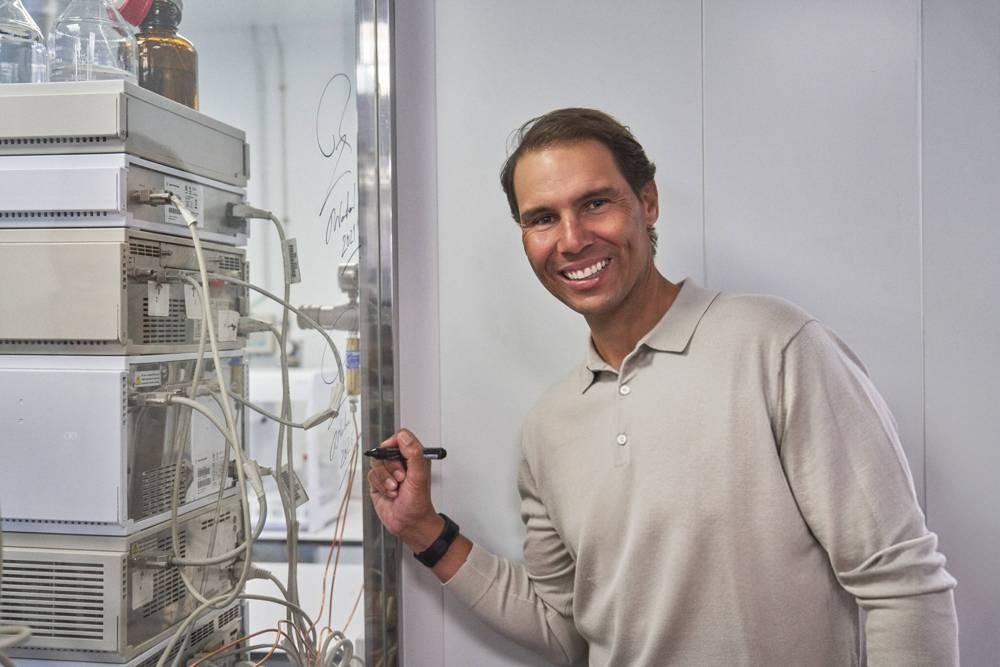 A smiling man in a beige sweater signs a glass panel beside stacked laboratory equipment with tubing, in a clean lab setting.