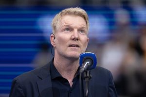 A man with short blond hair and a dark navy jacket speaks into a blue microphone, wearing an earpiece, in front of a blurred blue arena backdrop.