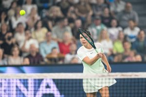 A tennis player in a white dress swings a dark racquet at a yellow ball during a match, with a crowd watching behind the court.