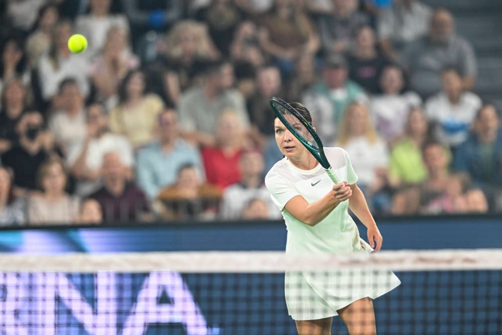 A tennis player in a white dress swings a dark racquet at a yellow ball during a match, with a crowd watching behind the court.