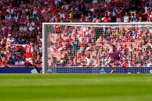Arsenal player number 11 in red and white shooting at goal as a pink-kitted goalkeeper dives, in a packed stadium