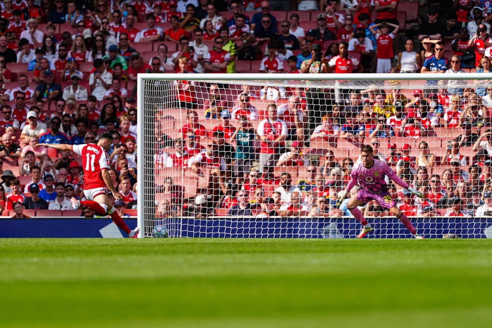 Arsenal player number 11 in red and white shooting at goal as a pink-kitted goalkeeper dives, in a packed stadium