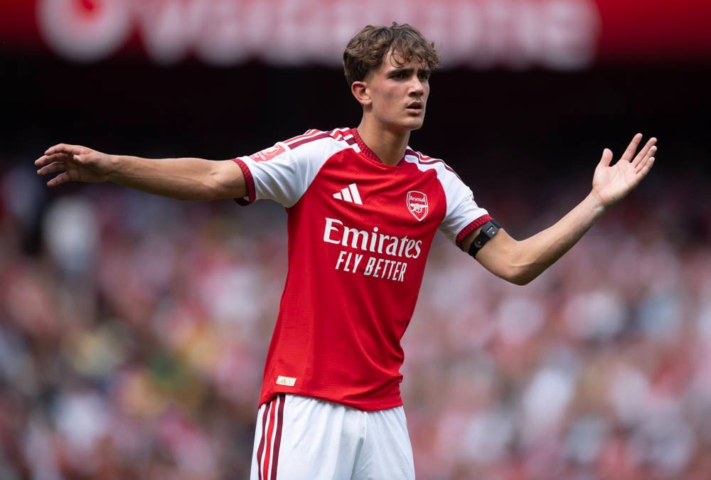 Young footballer in red and white Arsenal kit with Emirates sponsor, arms outstretched, speaking during a match at the stadium.