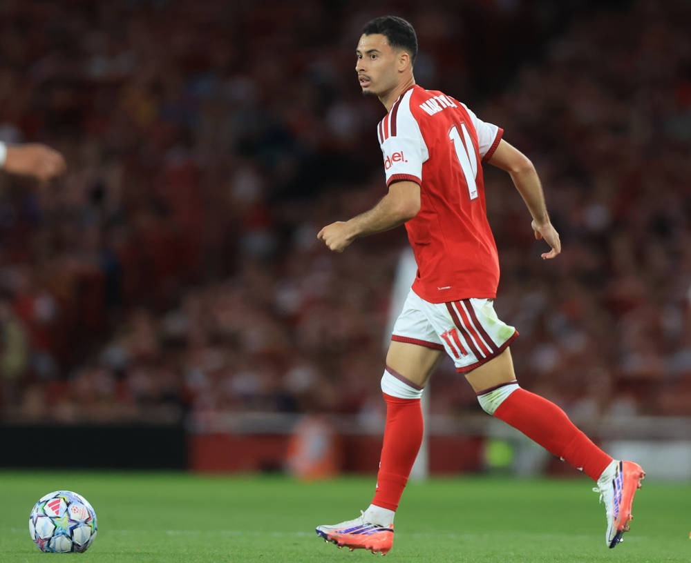 Football player in red Arsenal kit with number 11 running on the pitch, ball nearby, at a packed stadium. Name on shirt partly visible.