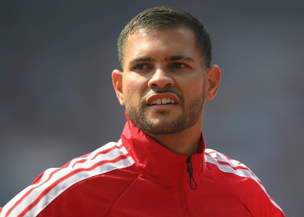 Man in red zip-up athletic jacket with white stripes, short hair and stubble, looking to the side at an outdoor stadium setting
