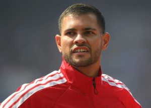 Man in red zip-up athletic jacket with white stripes, short hair and stubble, looking to the side at an outdoor stadium setting