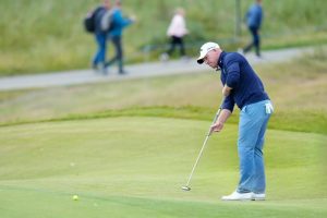 Golfer in navy sweater and white cap putts on a green while spectators walk on a path behind him at a tournament