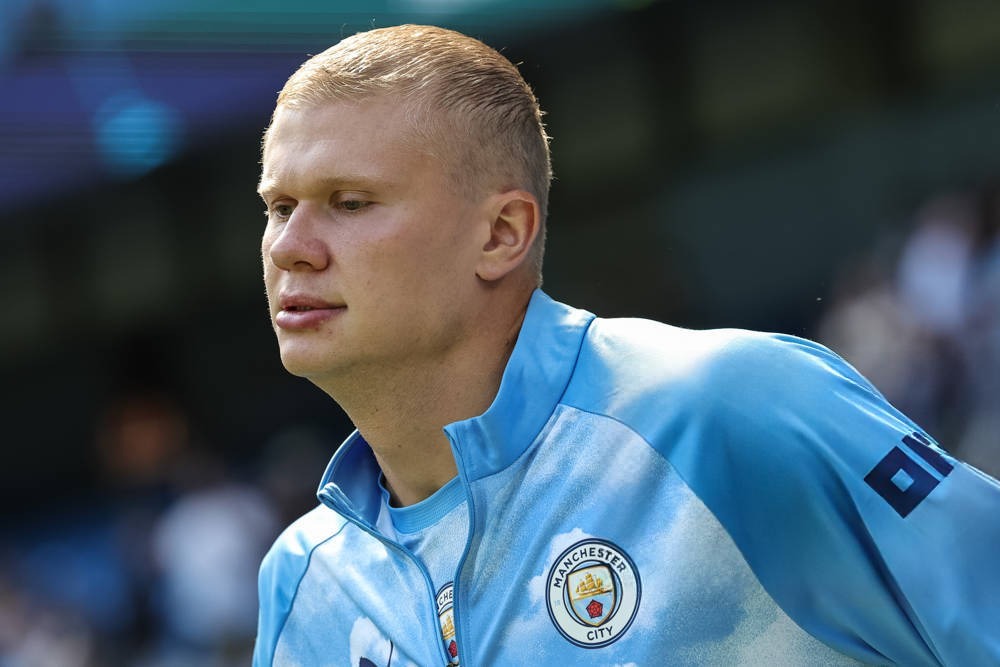 A blond young man in a light blue Manchester City jacket with the club crest, looking to the side at a stadium.