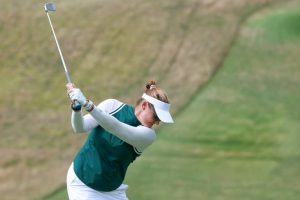 Golfer in green top, white visor and white sleeves mid-swing with an iron on a grassy course